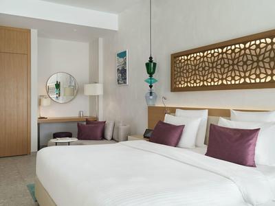 Helles Hotelzimmer mit großem Bett, violetten Kissen und dekorativer Wandbeleuchtung.