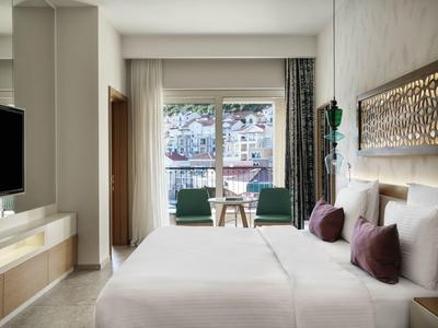 Modernes Hotelzimmer mit großem Doppelbett, zwei grünen Sesseln und Balkon mit Blick auf Stadt.