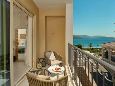 Balkon eines Hotelzimmers mit Sitzbereich, Blick auf Meer und Berge bei sonnigem Wetter.
