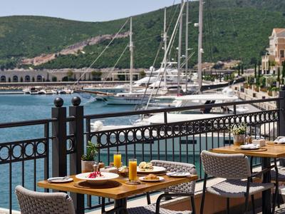Terrassenrestaurant mit gedeckten Tischen und Blick auf Jachten im Hafen vor grünen Hügeln.