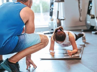 Frau macht Plank-Übung auf Matte, während Trainer sie in modernem Fitnessstudio beobachtet.