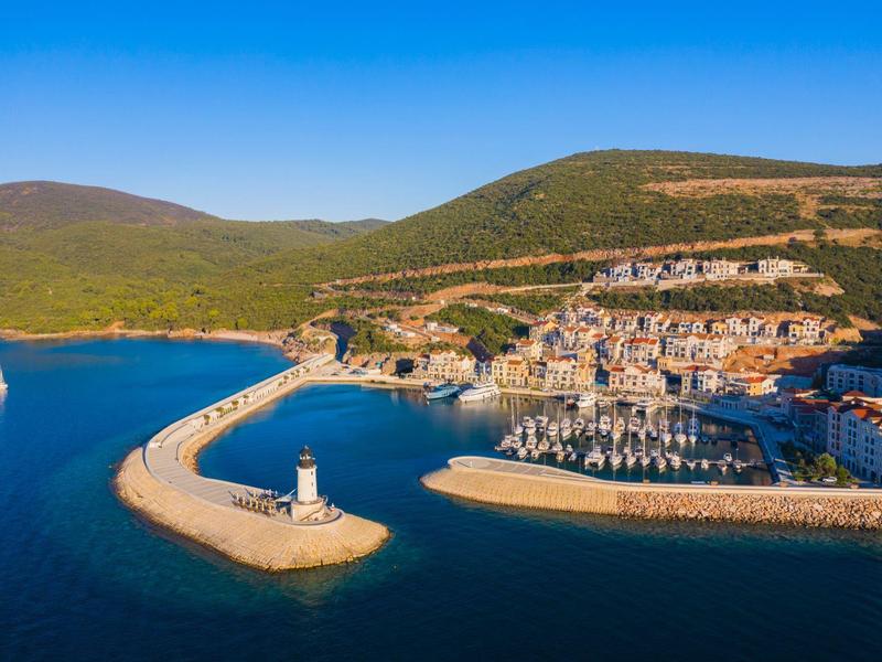 Haven met vuurtoren en jachten bij een kustplaats met heuvels op de achtergrond.
