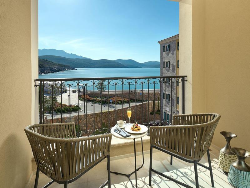 Een balkon met twee stoelen en een tafel met drankjes met uitzicht op zee en bergen.