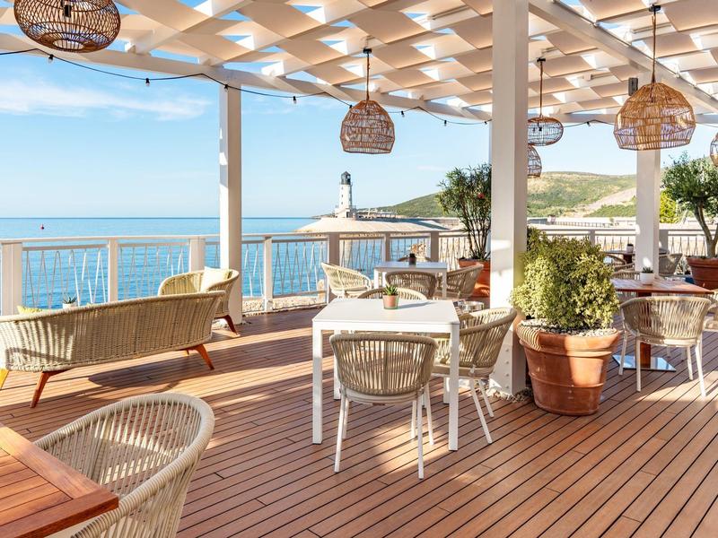 Terras met houten vloer, witte tafels en stoelen met uitzicht op zee en kustlijn.
