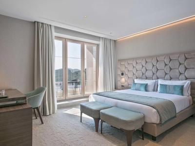 Modernes Schlafzimmer mit großem Bett, gepolstertem Kopfteil, zwei Sitzbänken und Balkonfenstern.