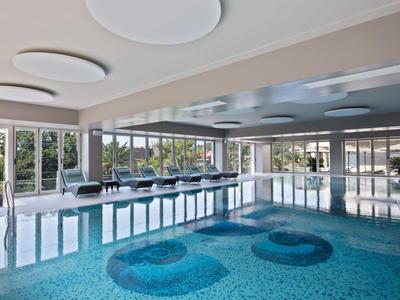 Jasny basen kryty z niebieską mozaiką, dużymi oknami i nowoczesnym oświetleniem sufitowym.