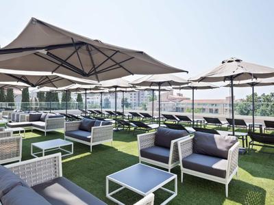 Taras na dachu z białymi meblami wypoczynkowymi, parasolami i zieloną sztuczną trawą pod czystym niebem.