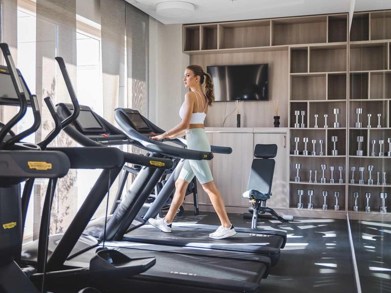 Frau läuft auf Laufband in hellem Fitnessraum mit Regalen voller Hanteln und großem Fenster.
