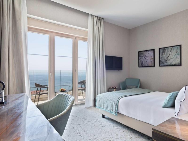 Helles Schlafzimmer mit großem Bett, Meerblick, zwei Stühlen, Schreibtisch und beiger Wanddekoration.