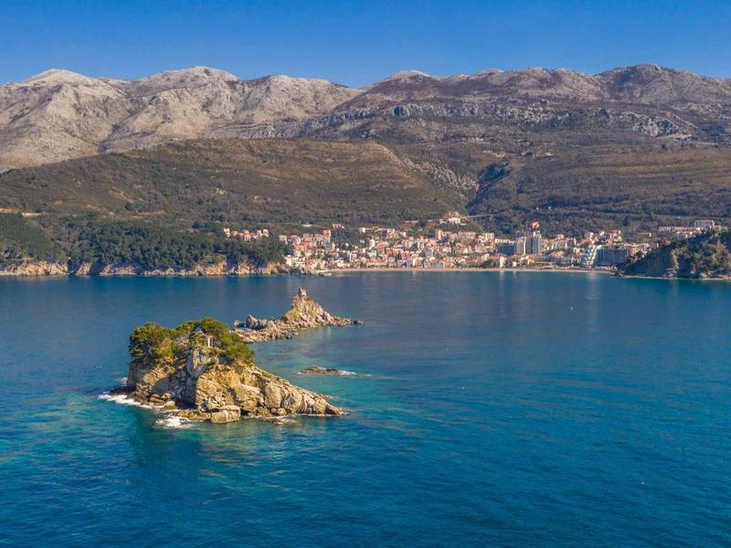 Kleine Insel mit Vegetation vor Küstenstadt und Gebirgslandschaft unter klarem blauen Himmel.