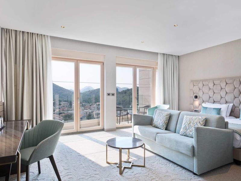 Heller Wohnbereich mit Sofas, Glastisch, Schreibtisch, Stuhl und großem Fenster mit Bergblick.