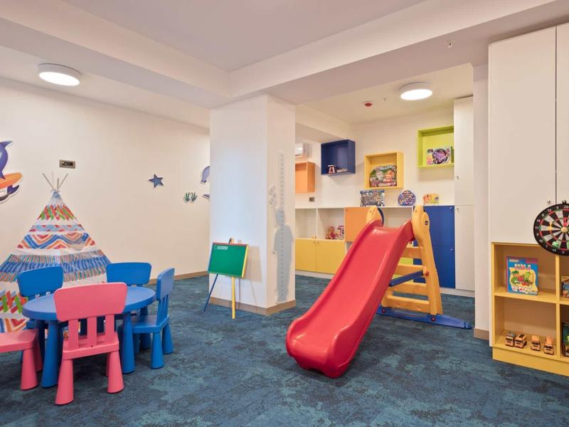 Kinderspielraum mit bunten Tischen, Stühlen, Rutsche, Bücherregal und Wanddekorationen.