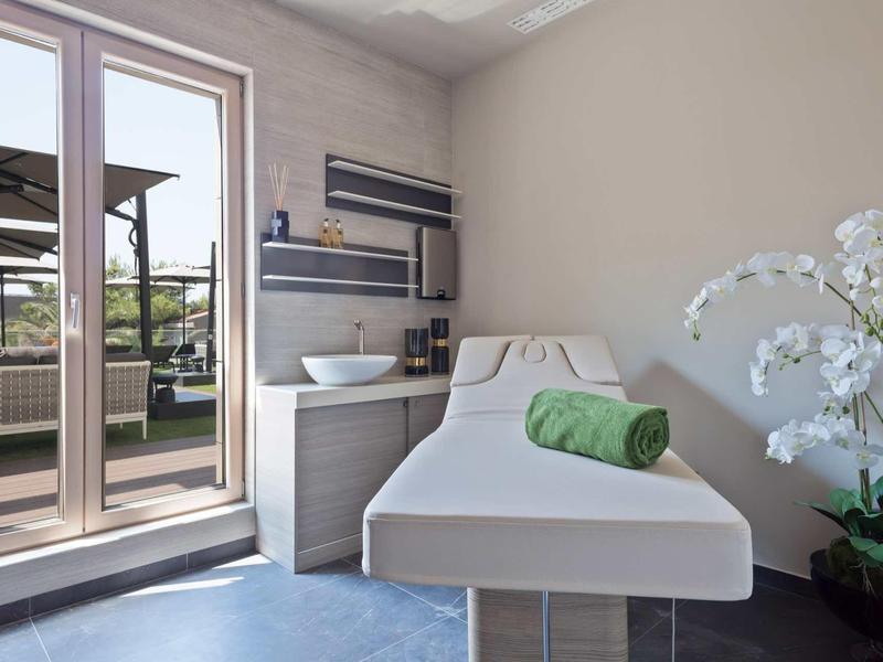 Heller Behandlungsraum mit Liege, grünem Handtuch, Orchidee und Zugang zu Terrasse mit Pool.