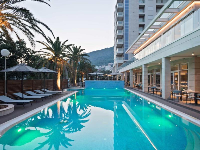 Moderner Außenpool bei Gebäude mit Glasfassade, umgeben von Palmen und Lounge-Bereichen.