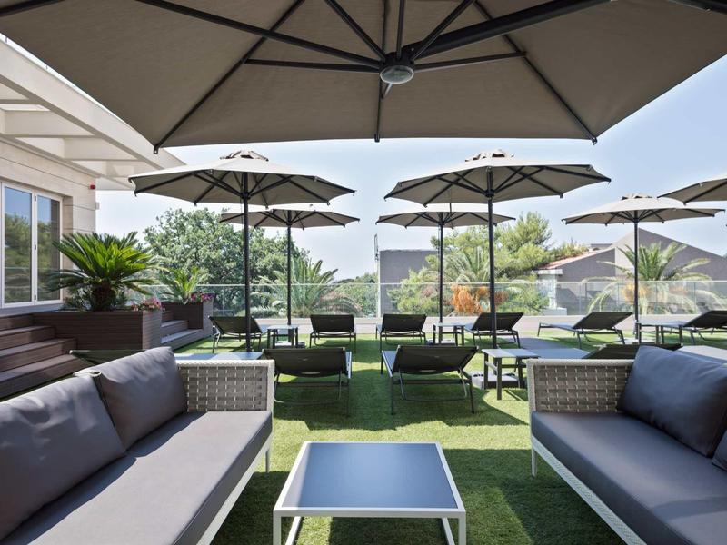 Terrasse mit grauen Sofas, Tisch und großen Sonnenschirmen auf grünem Kunstrasen bei klarem Himmel.