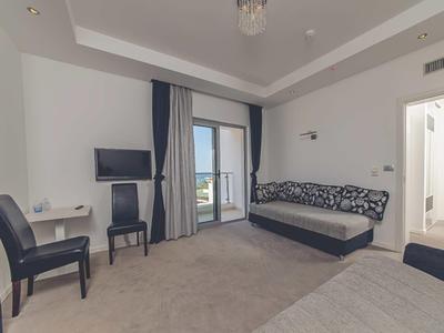 Hell eingerichtetes Zimmer mit grauem Teppich, Sofa, Stühlen, Fernseher und Balkon mit Meerblick.