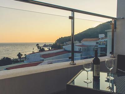 Sonnenuntergang über dem Meer mit Häusern, Weinflasche und zwei Gläsern auf Balkon.