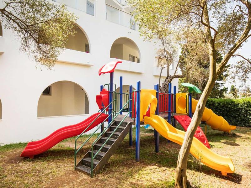 Bunter Kinderspielplatz mit Rutschen und Klettergerüst vor weißem Wohnhaus und Bäumen.