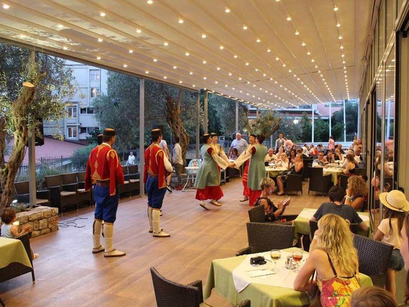 Menschen in bunter Tracht tanzen auf einer Bühne vor sitzendem Publikum unter Lichterhimmel.