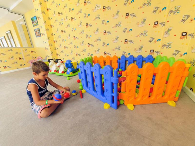Ein Junge spielt mit bunten Bällen vor einem kleinen, farbenfrohen Plastikzaun in einem hellen Raum.