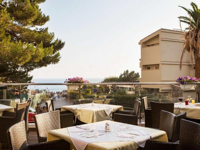 Terrasse mit gedeckten Tischen, Rattanstühlen und Blick auf Meer und Palmen im Hintergrund.