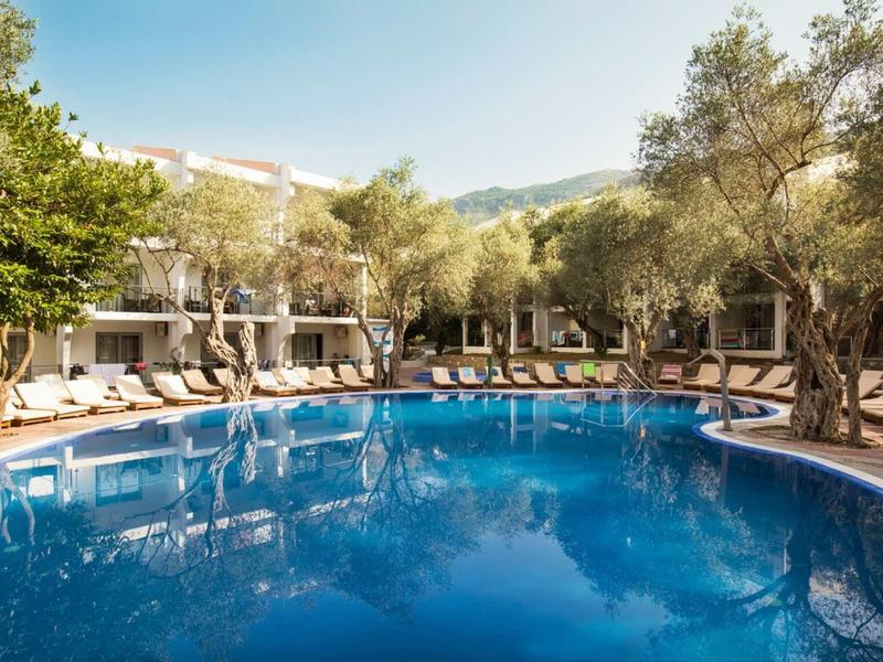 Runder Pool mit blauer Wasserfläche, umgeben von Liegestühlen und Bäumen vor einem weißen Hotel.