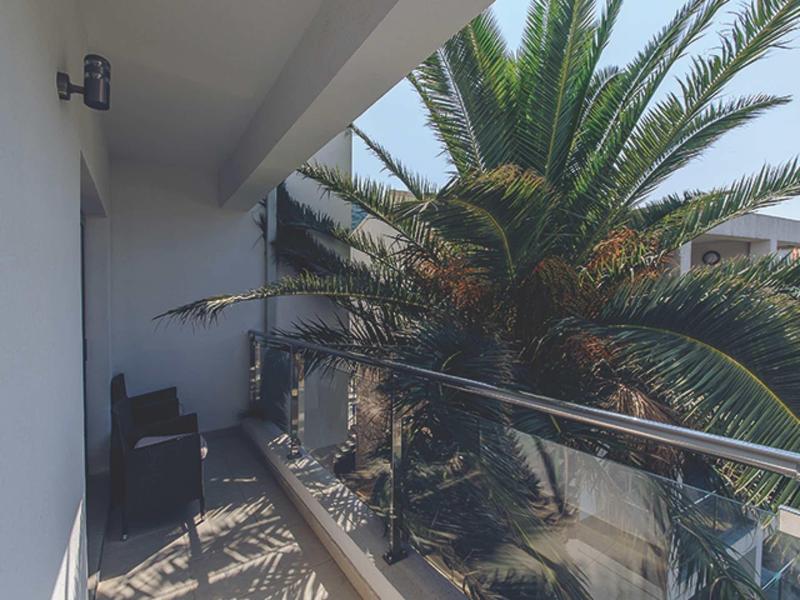 Balkon mit Glasgeländer, zwei Stühlen und großer Palme, helle Hauswand und blauer Himmel.