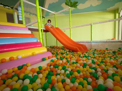Kind rutscht auf orangener Rutsche in bunt gefülltes Bällebad mit bunten Stufen und Netzumzäunung.