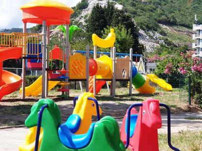 Bunter Spielplatz mit Rutsche, Schaukeln und Klettergerüst vor grüner Berglandschaft.