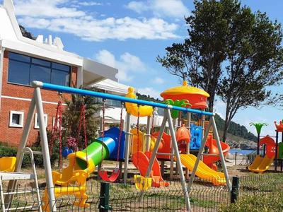 Bunte Spielplatz-Geräte aus Metall und Kunststoff auf grünem Rasen vor rotem Haus und blauem Himmel.