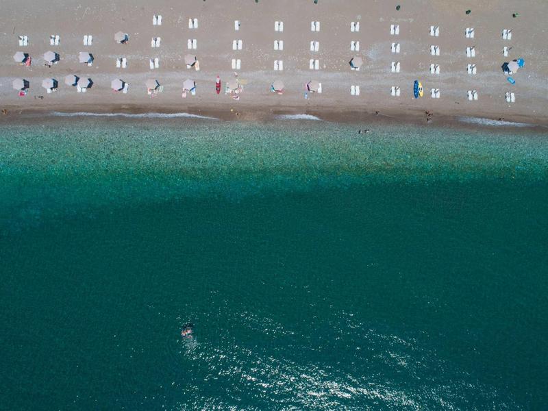 Luftaufnahme eines Strandes mit geordneten Sonnenschirmen, blau-grünem Wasser und feinem Sand.