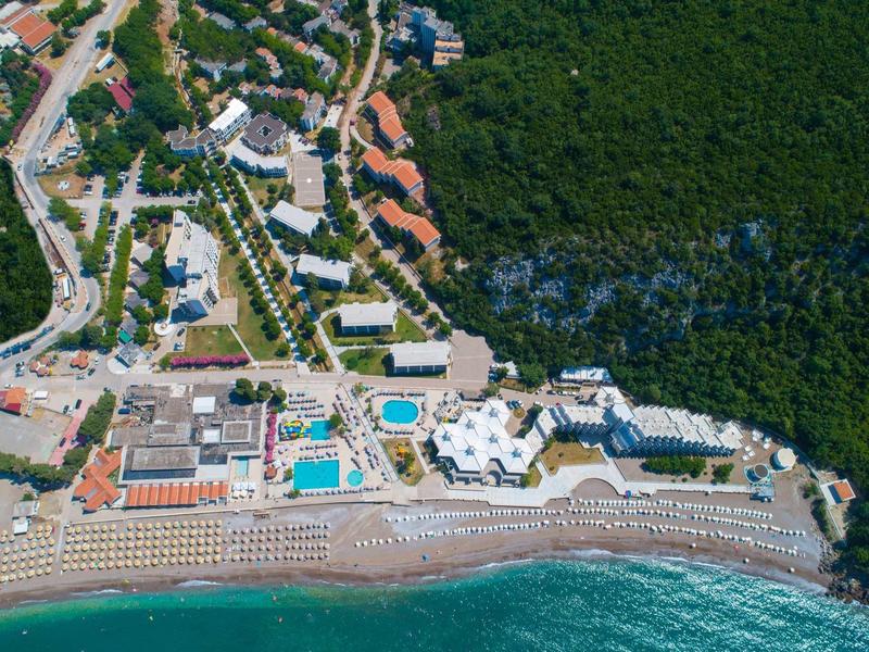 Luftaufnahme eines Küstenresorts mit Pool, Strand voller Liegestühle und dichtem Wald im Hintergrund.