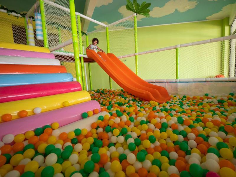 Kind rutscht auf orangener Rutsche in bunt gefülltes Bällebad mit bunten Stufen und Netzumzäunung.