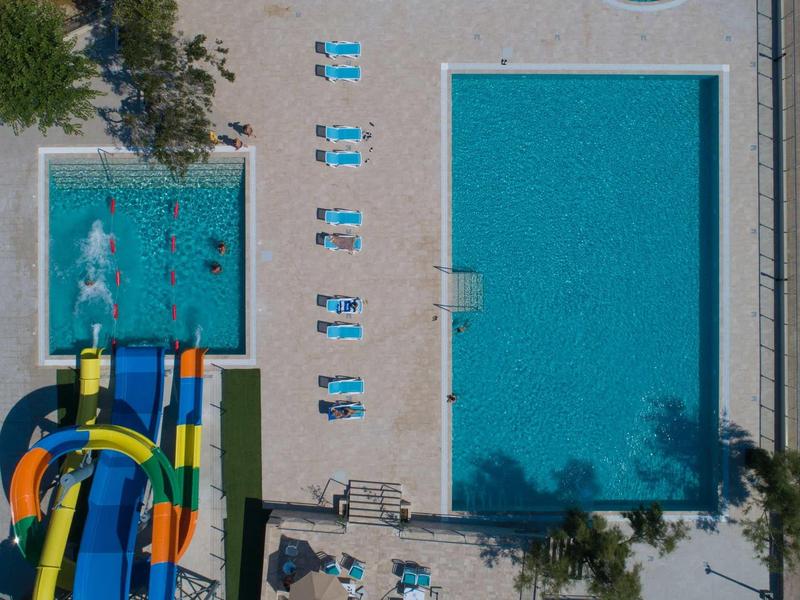 Vogelperspektive eines Schwimmbads mit Wasserrutschen, großem Becken, Kinderbecken und Liegestühlen.