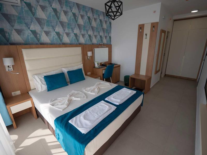 Hell eingerichtetes Hotelzimmer mit großem Bett, blauer Deko, Holzmöbeln und geometrischer Wandgestaltung.