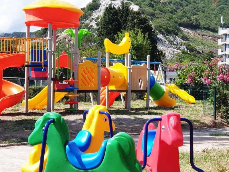 Bunter Spielplatz mit Rutsche, Schaukeln und Klettergerüst vor grüner Berglandschaft.
