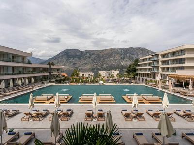 Großer Hotelpool mit Liegen und Sonnenschirmen vor Bergkulisse unter bewölktem Himmel.