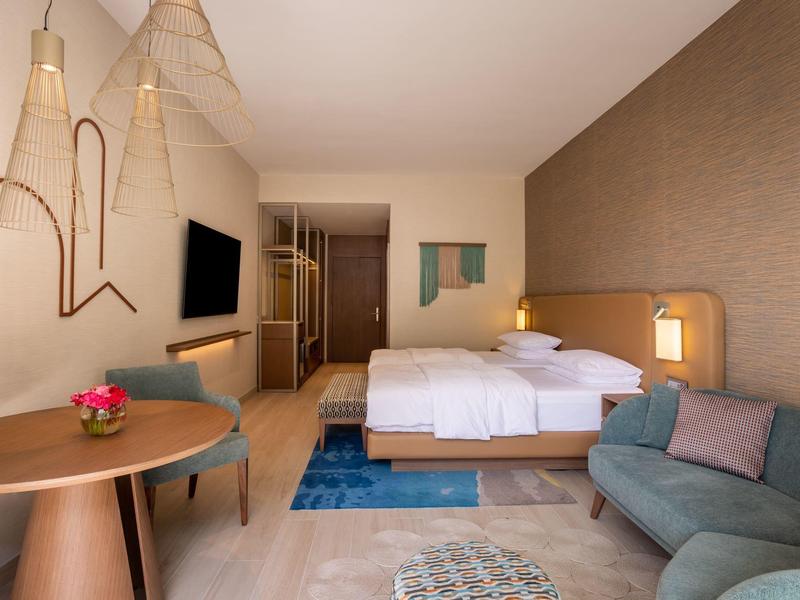 Moderní hotelový pokoj s manželskou postelí, posezením a kulatým dřevěným stolem v teplých tónech.