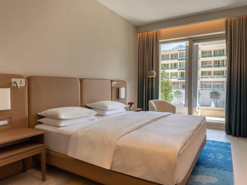 Moderní hotelový pokoj s manželskou postelí, nočními stolky a velkým oknem s výhledem ven.