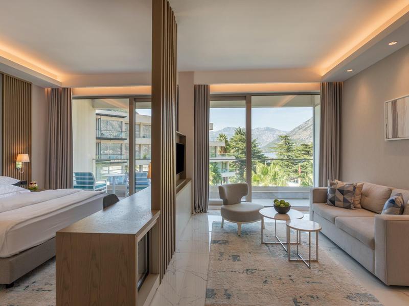 Moderní hotelový pokoj s postelí, pohovkou, křesly a velkými okny s výhledem na hory.