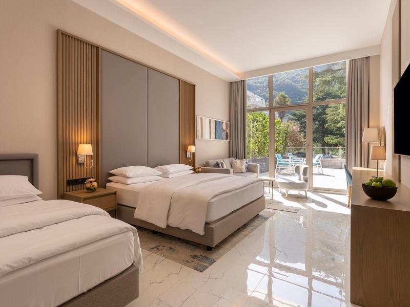 Světý hotelový pokoj se dvěma postelemi, moderním zařízením a balkonem s výhledem na hory.