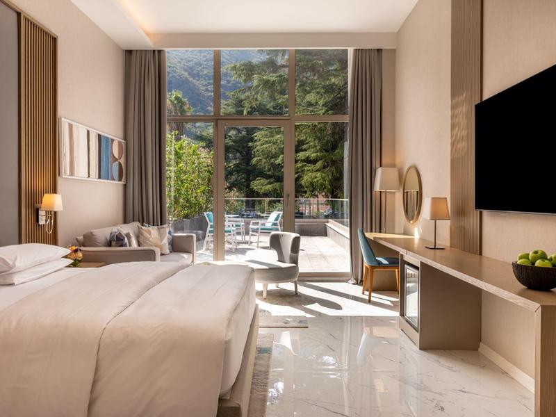 Světle hotelový pokoj s velkou postelí, psacím stolem, televizí a výhledem na terasu se zahradou.