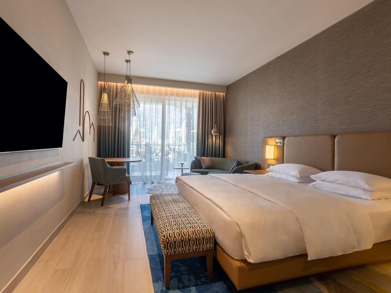 Moderní hotelový pokoj s manželskou postelí, televizí, křeslem a balkonem se závěsy.