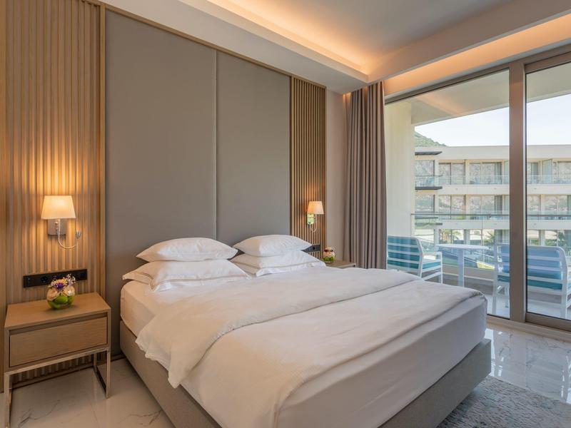 Moderní hotelový pokoj s velkou postelí, nočními stolky a balkonem s výhledem na budovu hotelu.