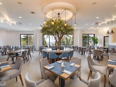 Světla hotelová restaurace s moderními stoly, židlemi a velkým stromem uprostřed.
