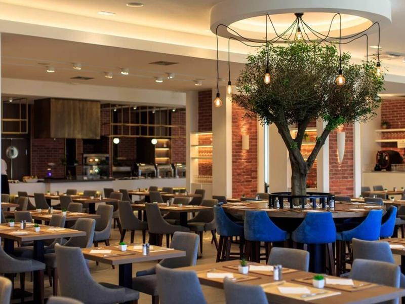 Modern hotel restaurant area with tables, chairs, and a decorative tree in the center.