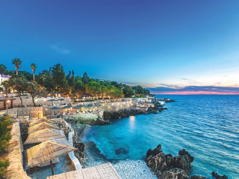 Beach resort with sun loungers and palm trees by clear blue sea at sunset.