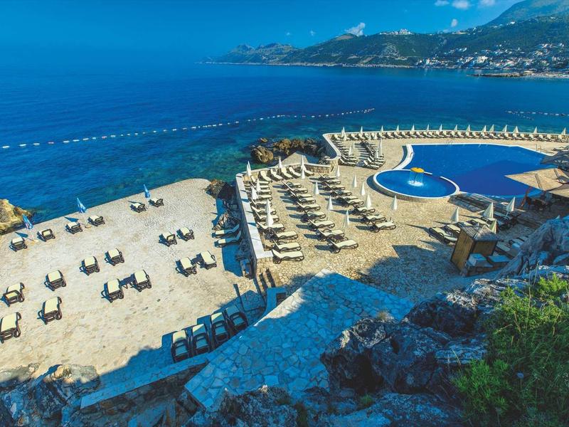 Aerial view of a beach resort with pool, sun loungers, and a view of the sea and mountains.