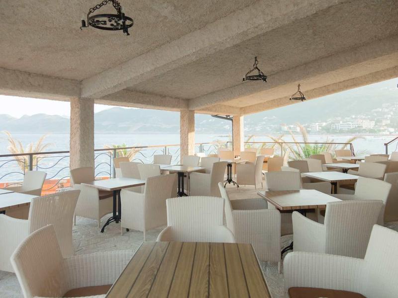 Covered terrace with white chairs and wooden tables overlooking the sea and mountains.