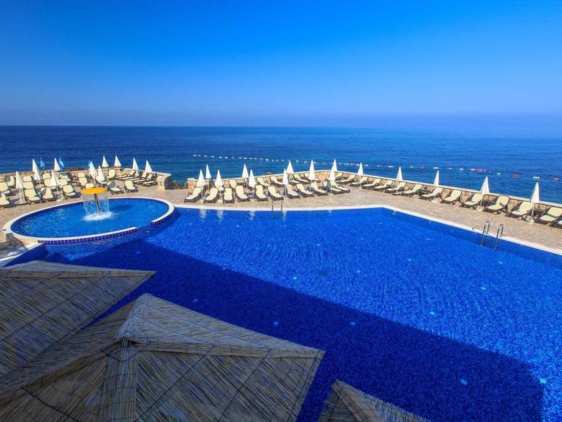 Large blue pool with lounge chairs and umbrellas by the sea under a clear sky.